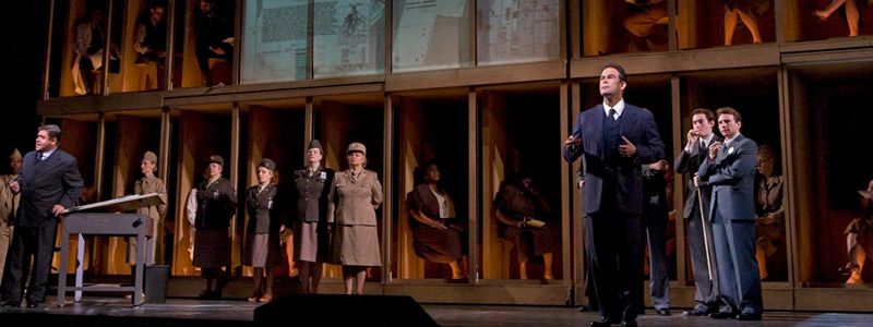 Theaterbühne mit zahlreichen Personen, teils Männer in dunklem Anzug, teils Frauen in Uniform, dahinter sitzen Personen in Kubikeln